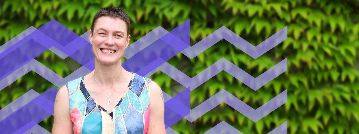 Smiling woman wearing multicolored top standing in front of bushes and a purple mountain pattern